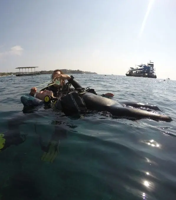 Instructor de buceo en Nusa Penida brindando respiración por octopus a un buzo que flota boca arriba en la superficie del mar cerca de embarcaciones.