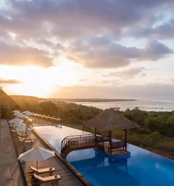 Atardecer en Nusa Penida sobre una piscina infinita con puente y gazebo, huéspedes relajándose en hamacas y sombrillas junto al mar.