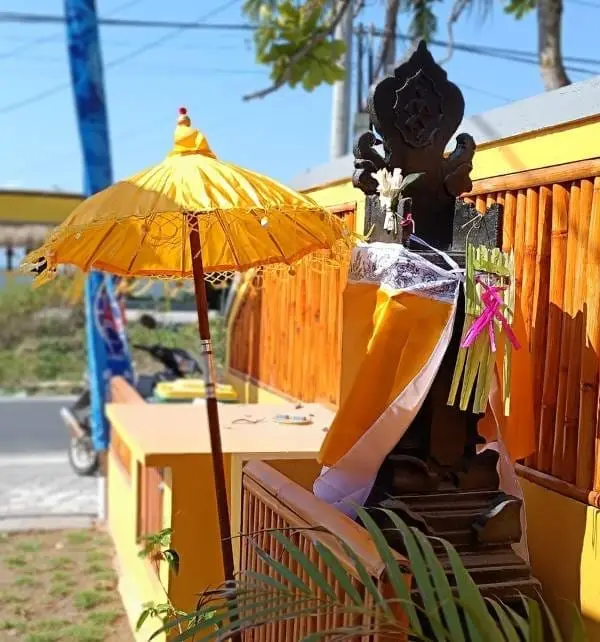 Altar balines decorado con tela naranja y ofrendas junto a una sombrilla amarilla en la calle de Nusa Penida