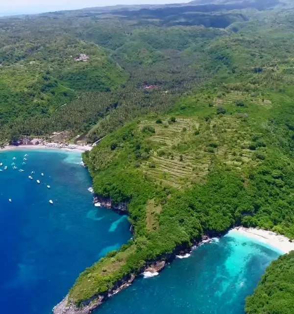 Aerial de Nusa Penida mostrando barcos en la bahía y turistas en playas de arena blanca entre acantilados y colinas verdes.