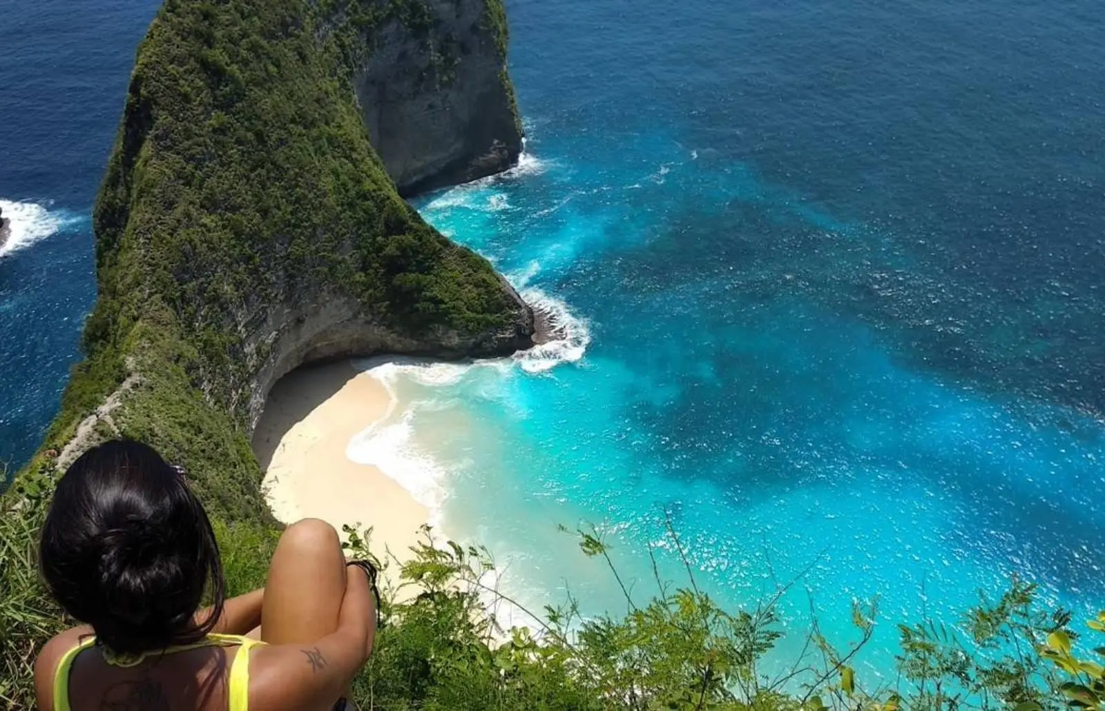 Mujer sentada en un acantilado mirando la playa de arena y el agua turquesa de Nusa Penida mientras las olas llegan a la bahía escondida.