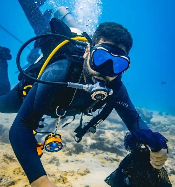 Buzo en Nusa Penida recogiendo basura del fondo marino con equipo de buceo y malla, escenas de limpieza submarina.