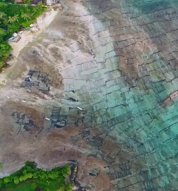 Vista aérea de Nusa Penida mostrando marea baja con redes y botes en la orilla y patrones de arrecife expuesto en el agua.