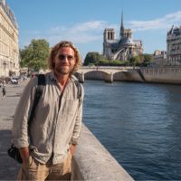 Hombre sonriente con barba y gafas de sol, mochila al hombro, apoyado en el muro junto al río Sena; al fondo la catedral de Notre-Dame.