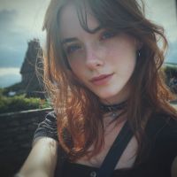 Joven con cabello castaño largo y flequillo, mirada directa y piel con pecas, retrato en exterior con cielo nublado y rocas difuminadas detrás.